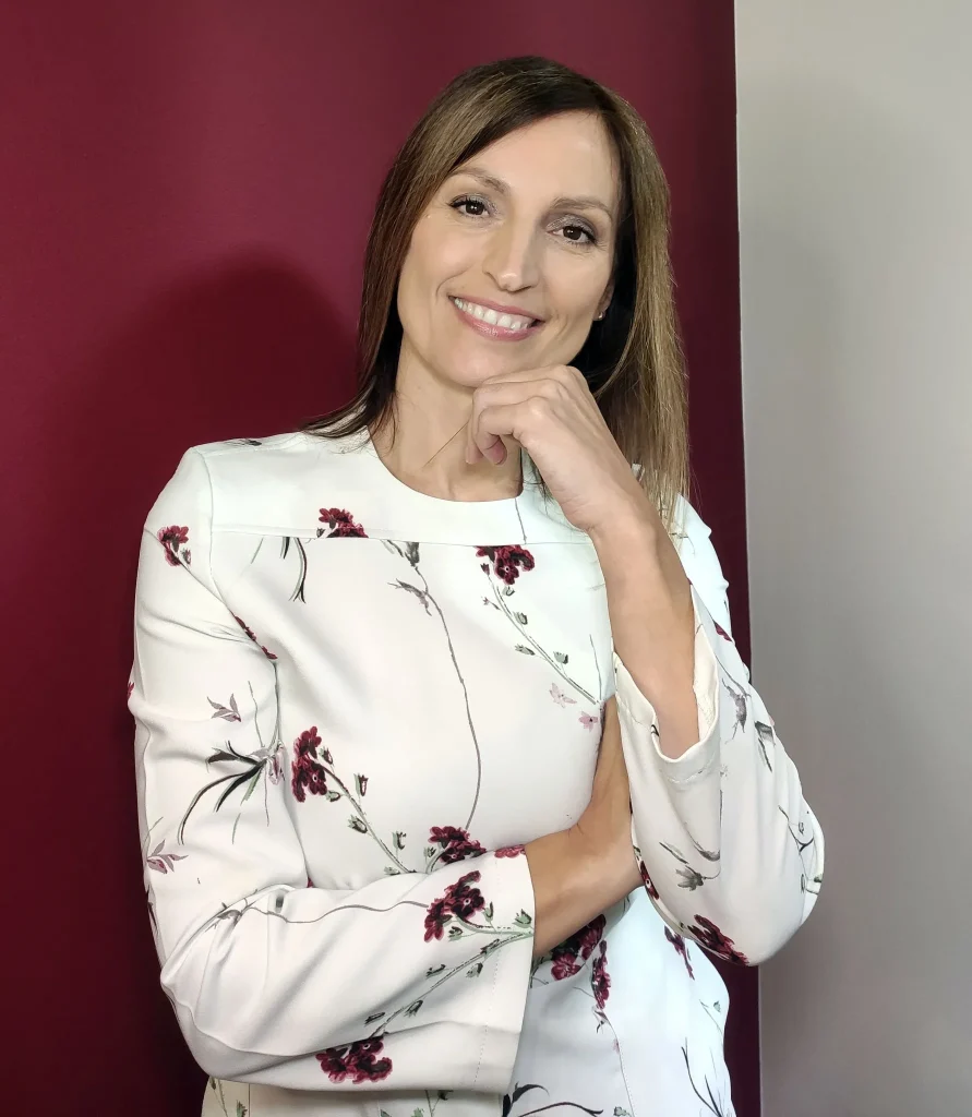 Olga Lestón con blusa blanca de estampado floral, sonriendo con expresión amigable y segura, con fondo en tonos burdeos y beige.