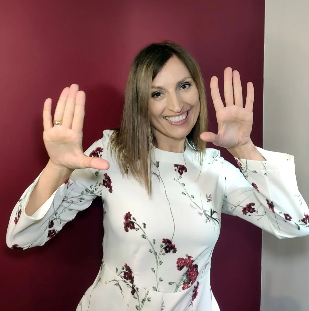 Olga Lestón con vestido blanco de flores rojas sonriendo y levantando las manos en un fondo granate y beige.