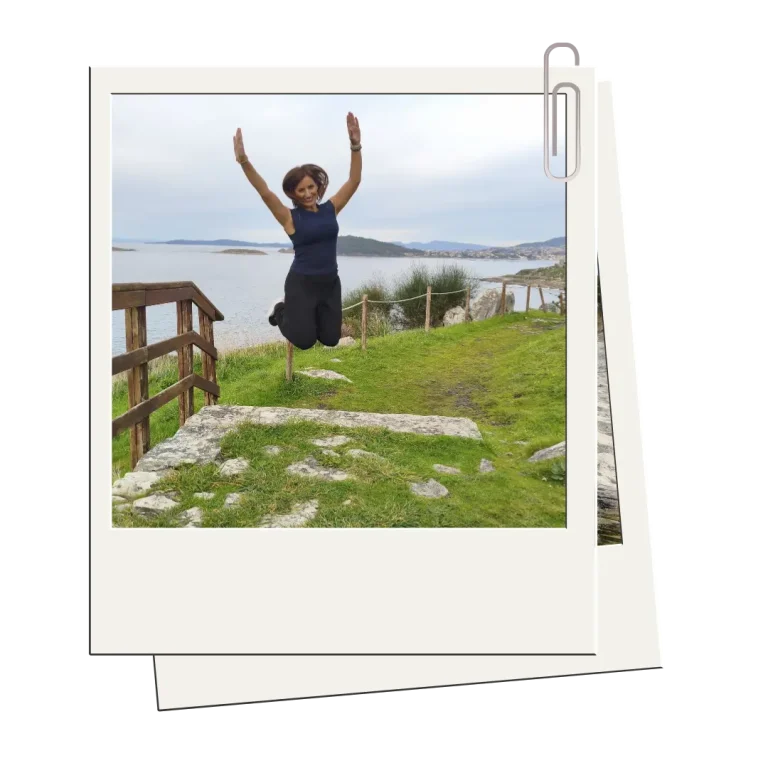 Mujer saltando con los brazos en alto sobre un sendero de piedra y césped, con vistas al mar y un paisaje natural de fondo.