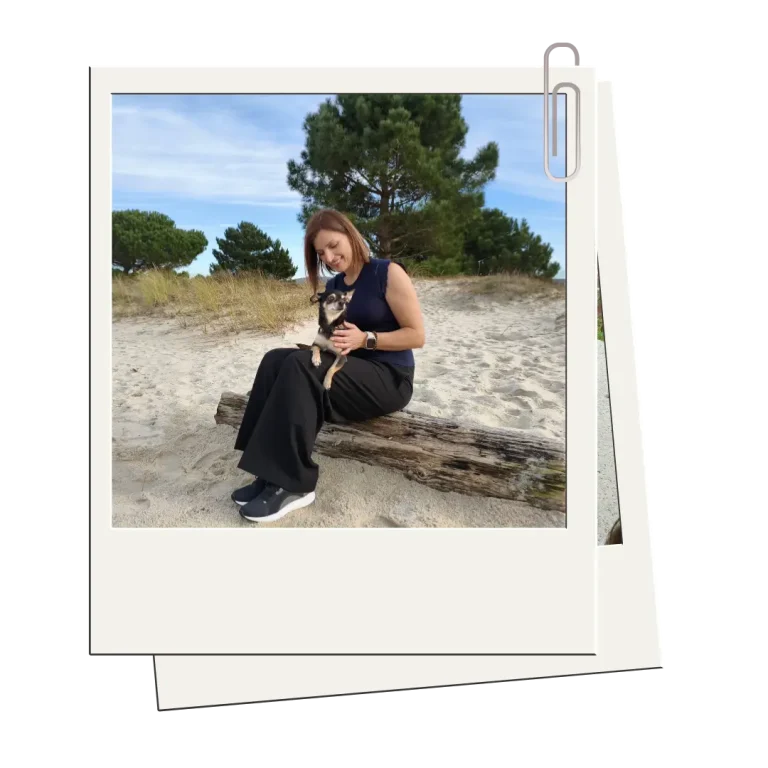 Mujer sentada en un tronco sobre la arena, sosteniendo a su pequeño perro mientras sonríe, con árboles y dunas al fondo.