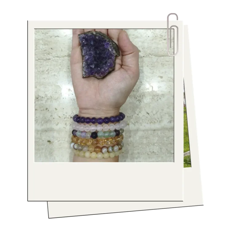 Mano sosteniendo una amatista en bruto, adornada con varias pulseras de piedras naturales de diferentes colores, sobre una superficie de mármol.
